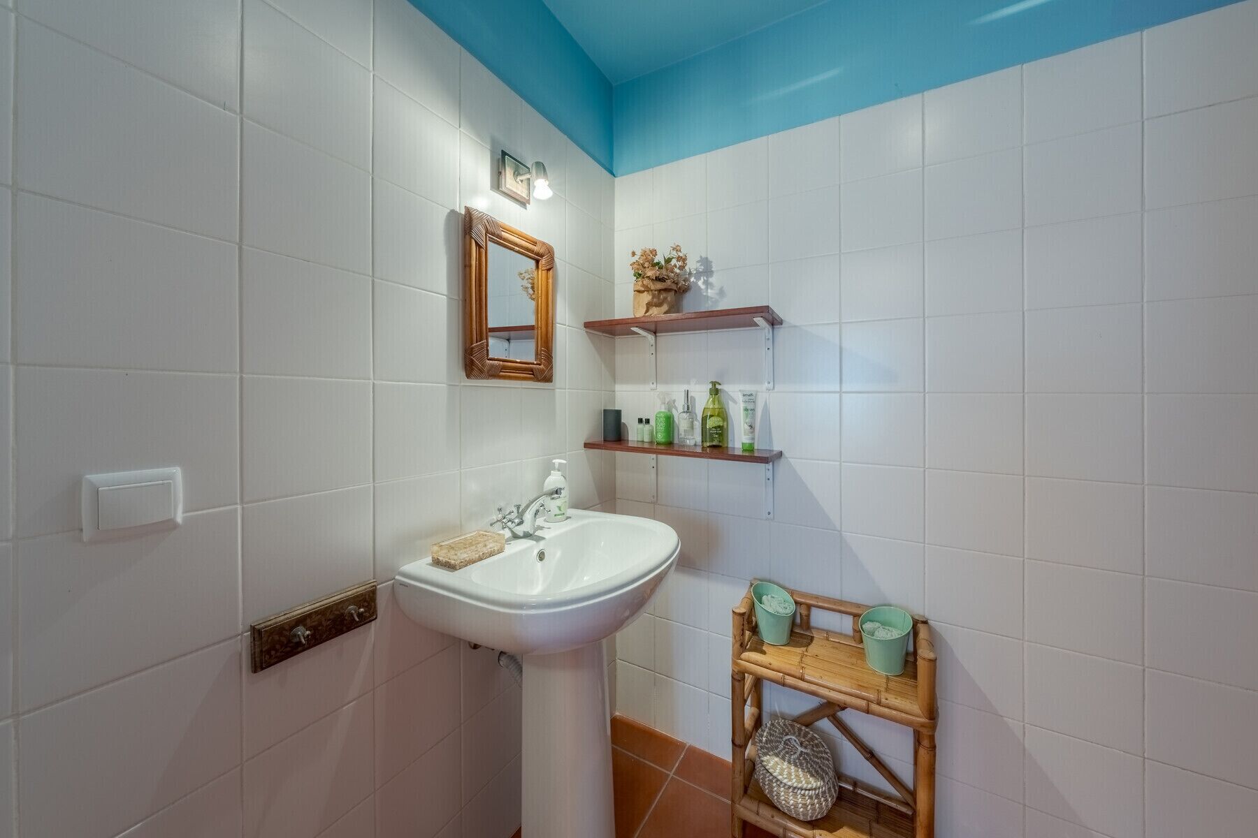 A bathroom with white tiled walls, a white sink, a wooden-framed mirror, and a shelf with toiletries.