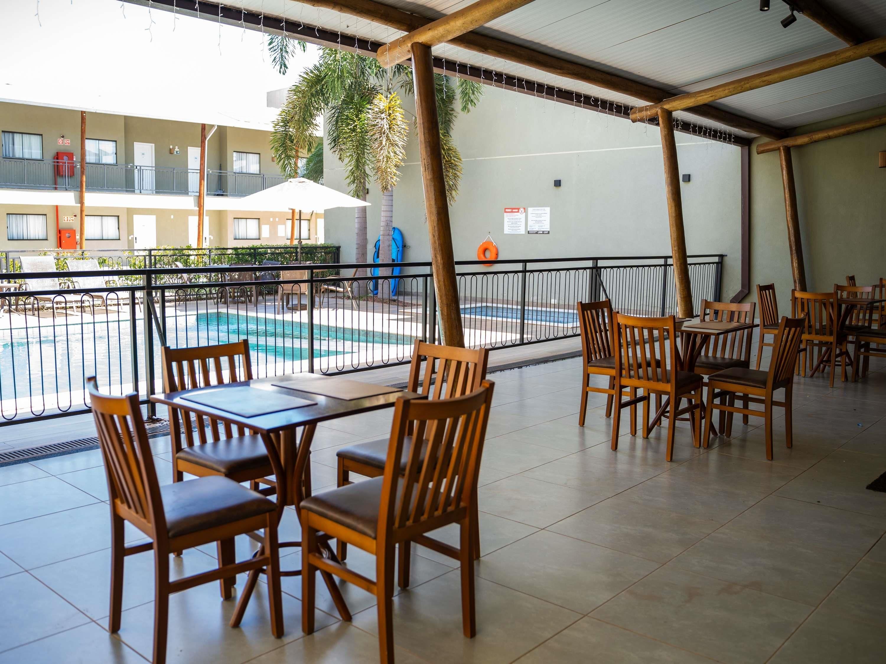A covered outdoor seating area with wooden tables and chairs, a pool in the background, and a building with balconies.