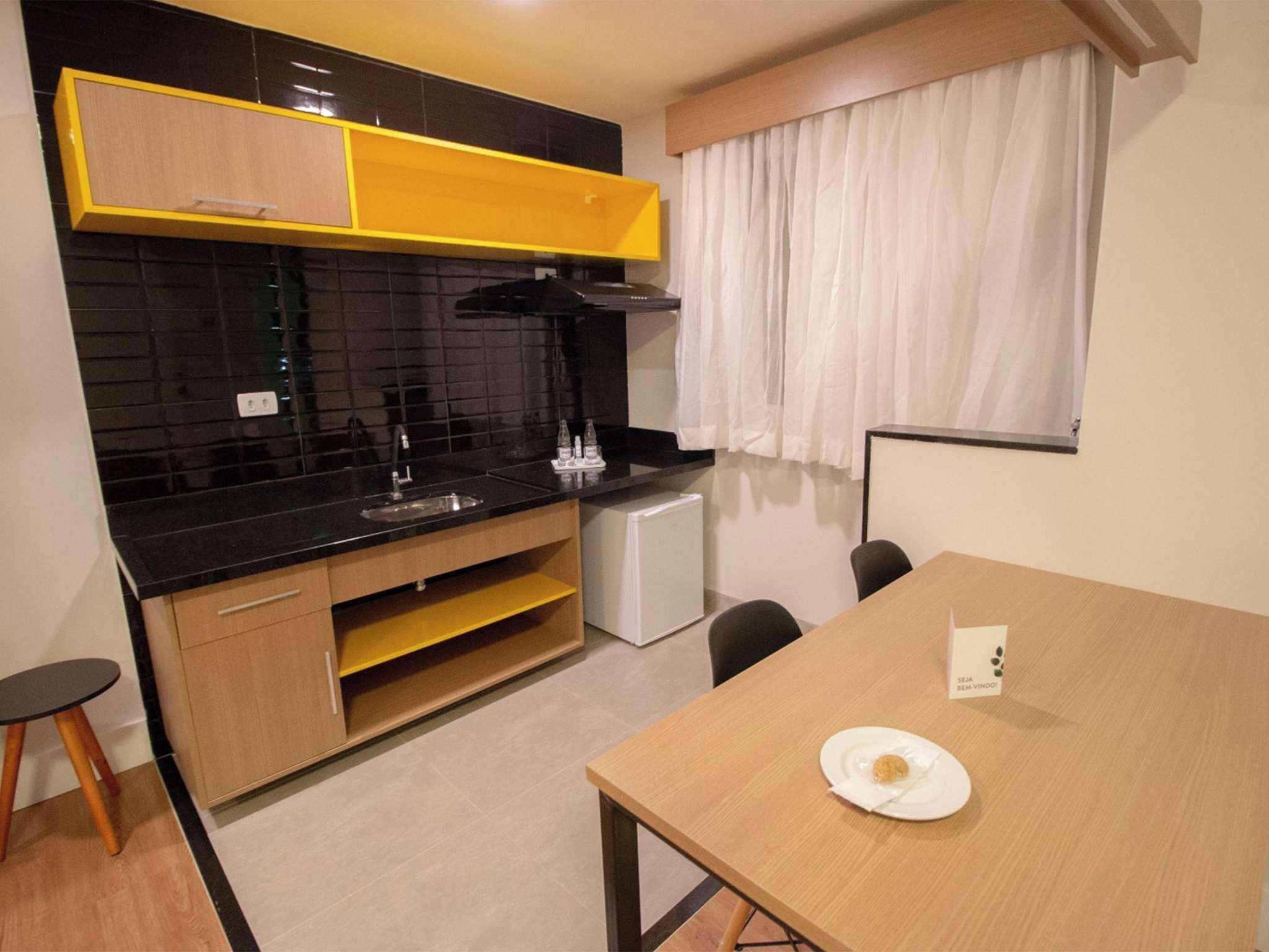 A compact kitchen with a black countertop, wooden cabinets, a small refrigerator, and a dining table with a plate.