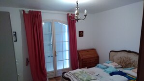 A bedroom with a bed, a wooden dresser, a chandelier, and a sliding glass door with red curtains. - Pretty villa on a hill on the edge of the forest (La Crau)