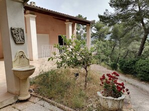 Ein Steinfbrunnen, ein kleiner Baum und ein Blumentopf in einer Gartenanlage, im Hintergrund ein Gebäude.