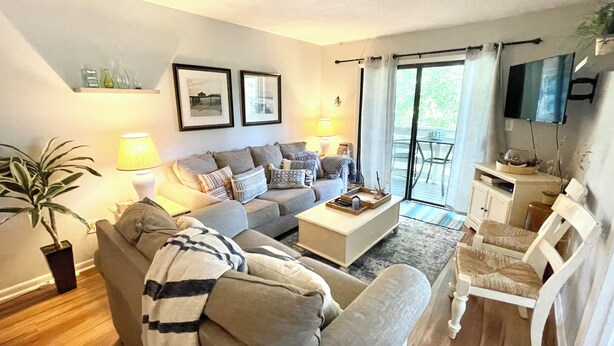Un salon comprenant un canapé, des fauteuils, une table basse et un téléviseur. On y trouve également une plante, une lampe et une fenêtre avec des rideaux.