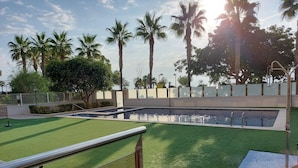 A swimming pool with a surrounding green lawn, palm trees, and a fence. - MARINA BEACH APARTMENT (Oropesa del Mar)