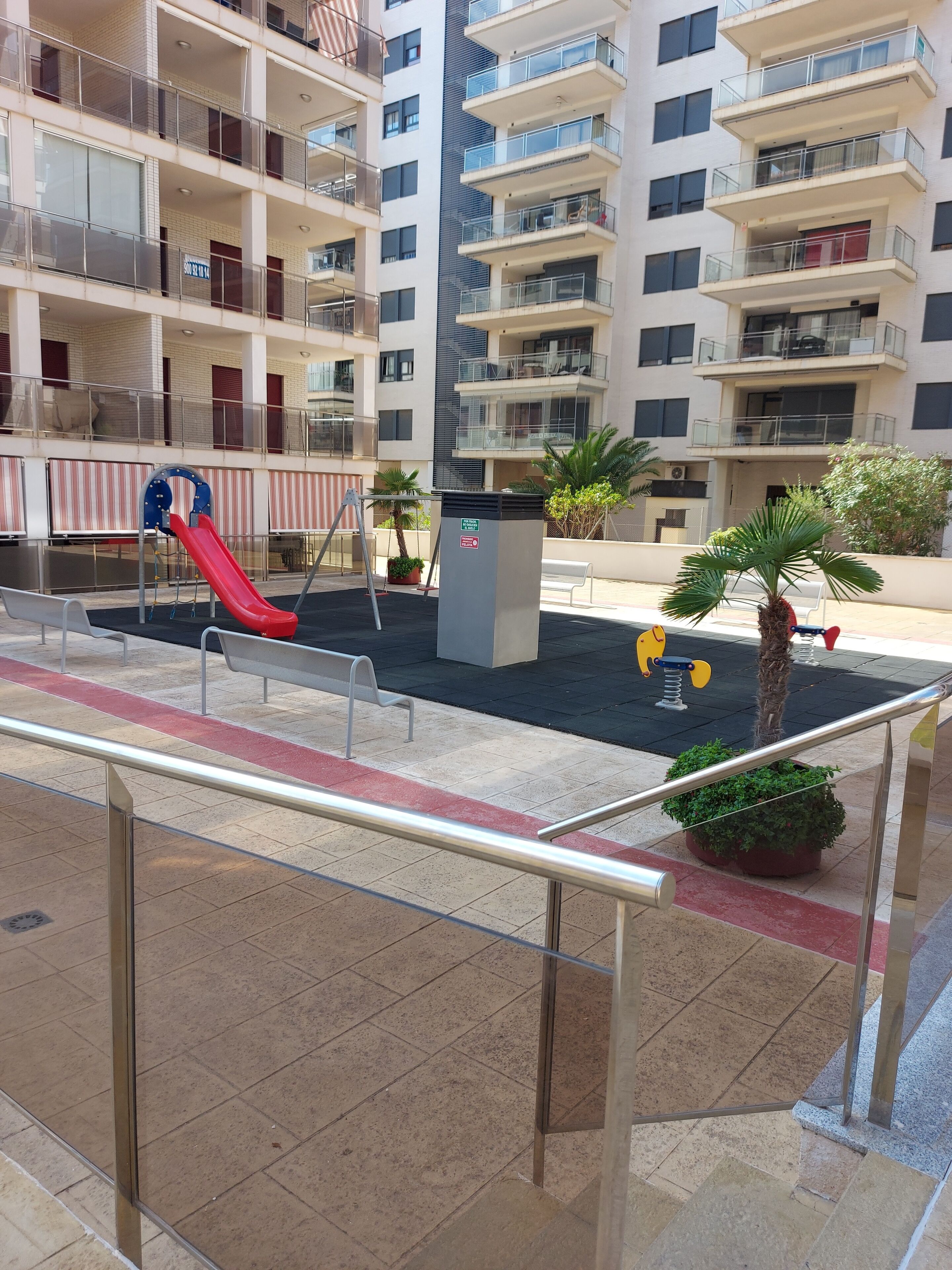 Un edificio residencial de varios pisos con balcones, un parque infantil y plantas en macetas.