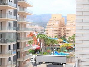 Un edificio de varios pisos con balcones, un parque acuático con toboganes y piscina, y un telón de fondo montañoso.