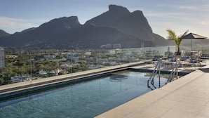 Una piscina en la azotea con agua azul cristalina, rodeada de sillones de descanso y una palmera, con vistas a un paisaje urbano y montañas.