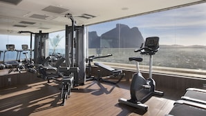 Un gimnasio con diverso equipamiento de ejercicio, amplias ventanas y vistas a un paisaje urbano.