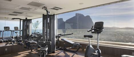 Un gimnasio con diverso equipamiento de ejercicio, amplias ventanas y vistas a un paisaje urbano.