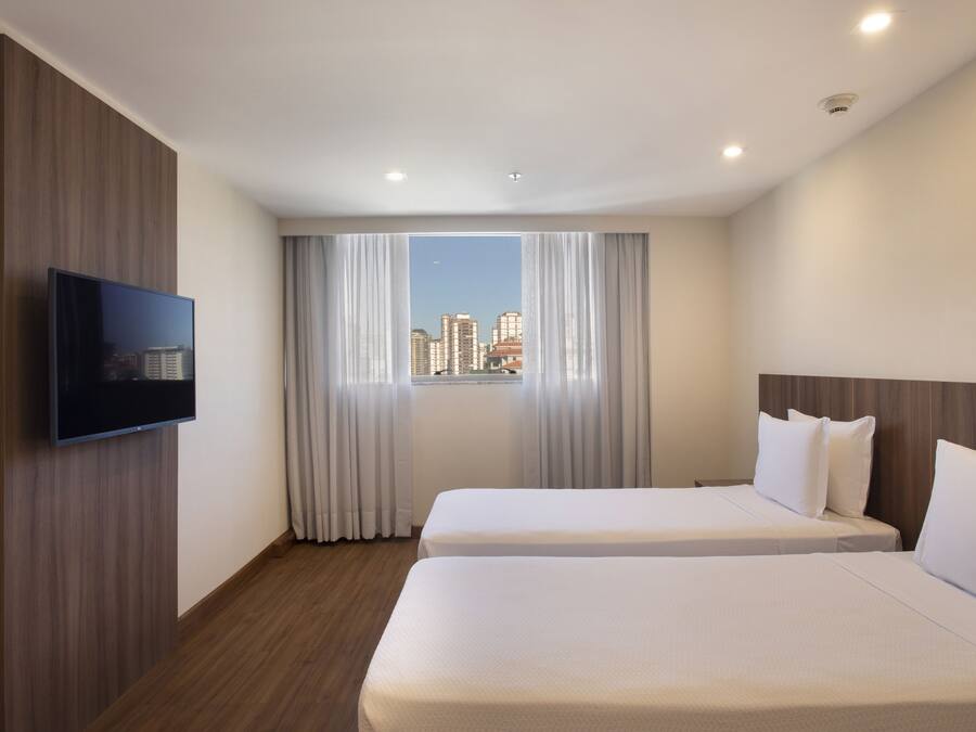 A hotel room with two beds, a flat-screen TV mounted on the wall, and a city view through the window.
