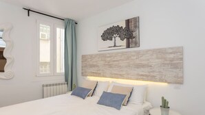 Une chambre à coucher comprenant un lit, une table de chevet, une fenêtre avec des rideaux, une œuvre d’art représentant un arbre accrochée au mur, et un radiateur.
