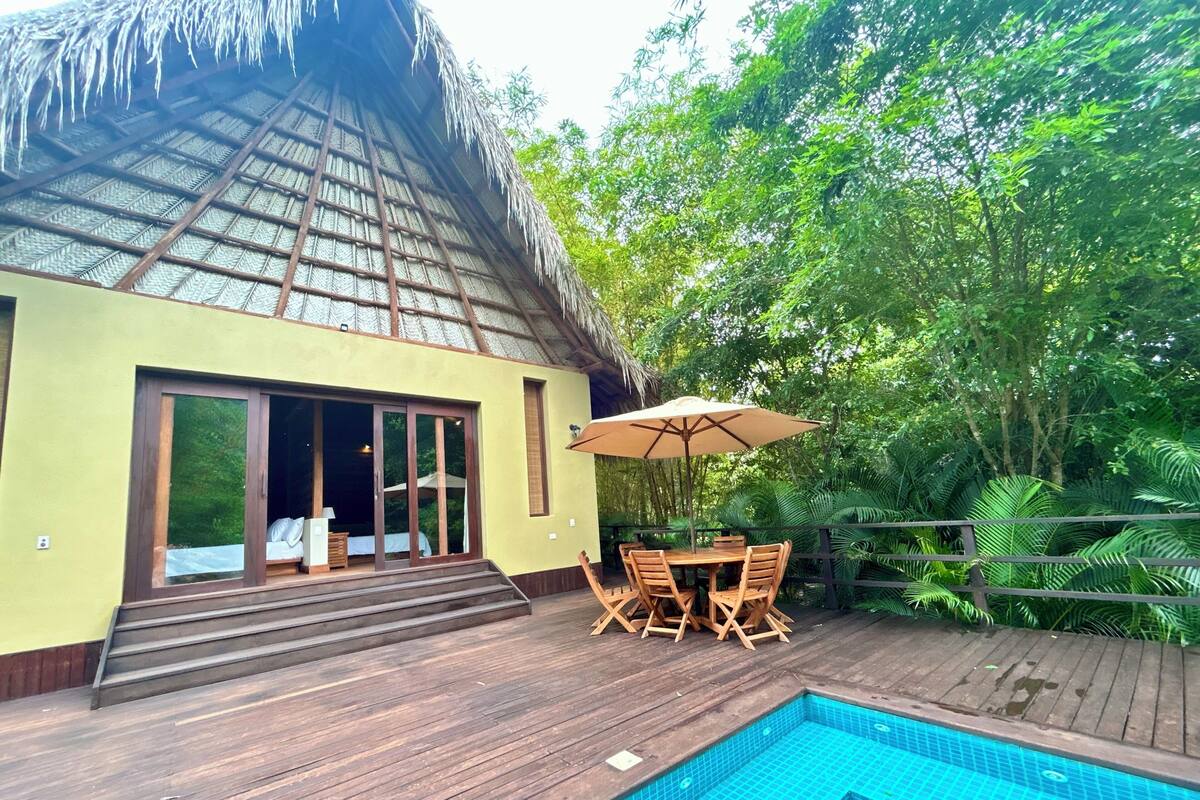 A poolside area with a wooden deck, a thatched-roof building, and a table with chairs.
