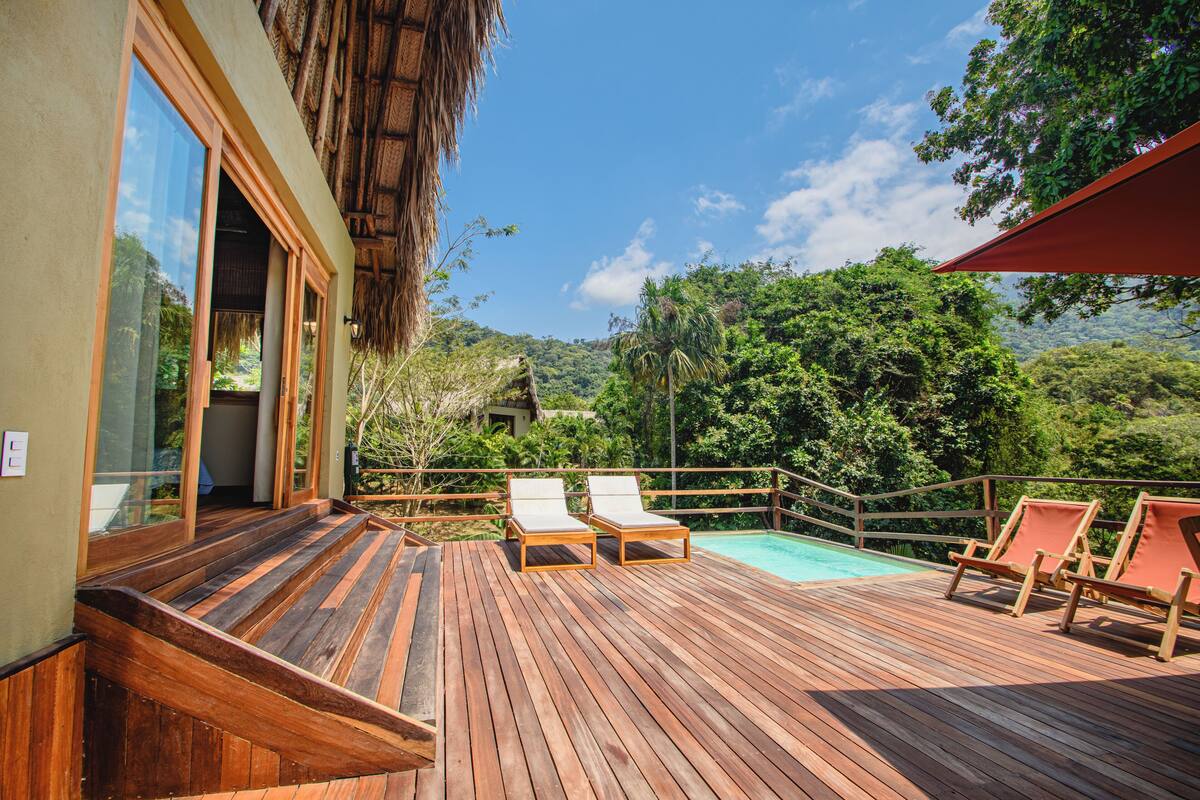A wooden deck with lounge chairs and a pool, surrounded by lush greenery and a mountain backdrop.