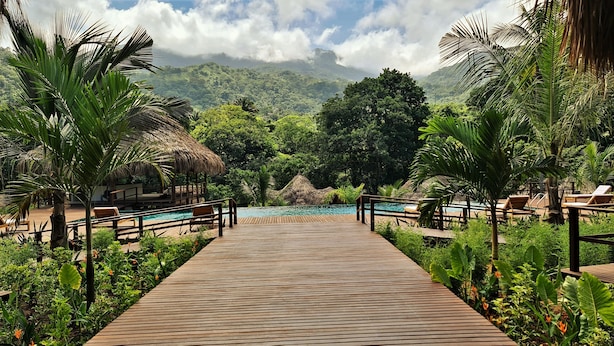 A wooden deck leading to a pool with lounge chairs and thatched-roof structures, surrounded by lush greenery and a mountainous backdrop. - Senda Watapuy (Santa Marta)