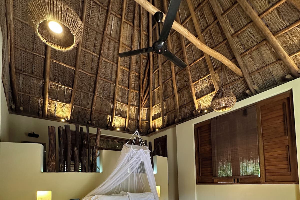 A room with a bed, a wooden bench, a ceiling fan, and a thatched roof.