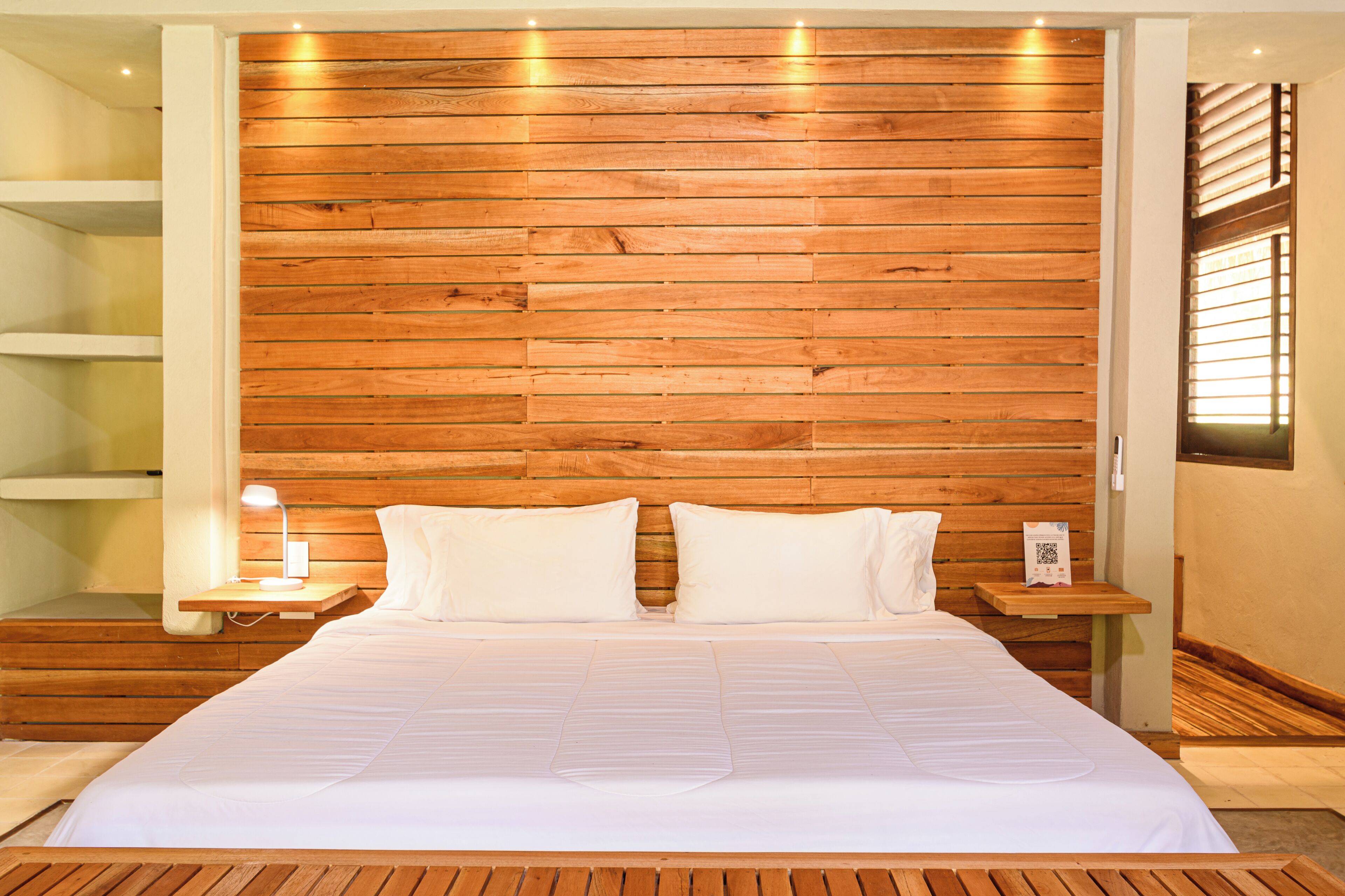 A hotel room with a wooden headboard, a large bed with white bedding, a bedside table with a lamp, and a small shelf.