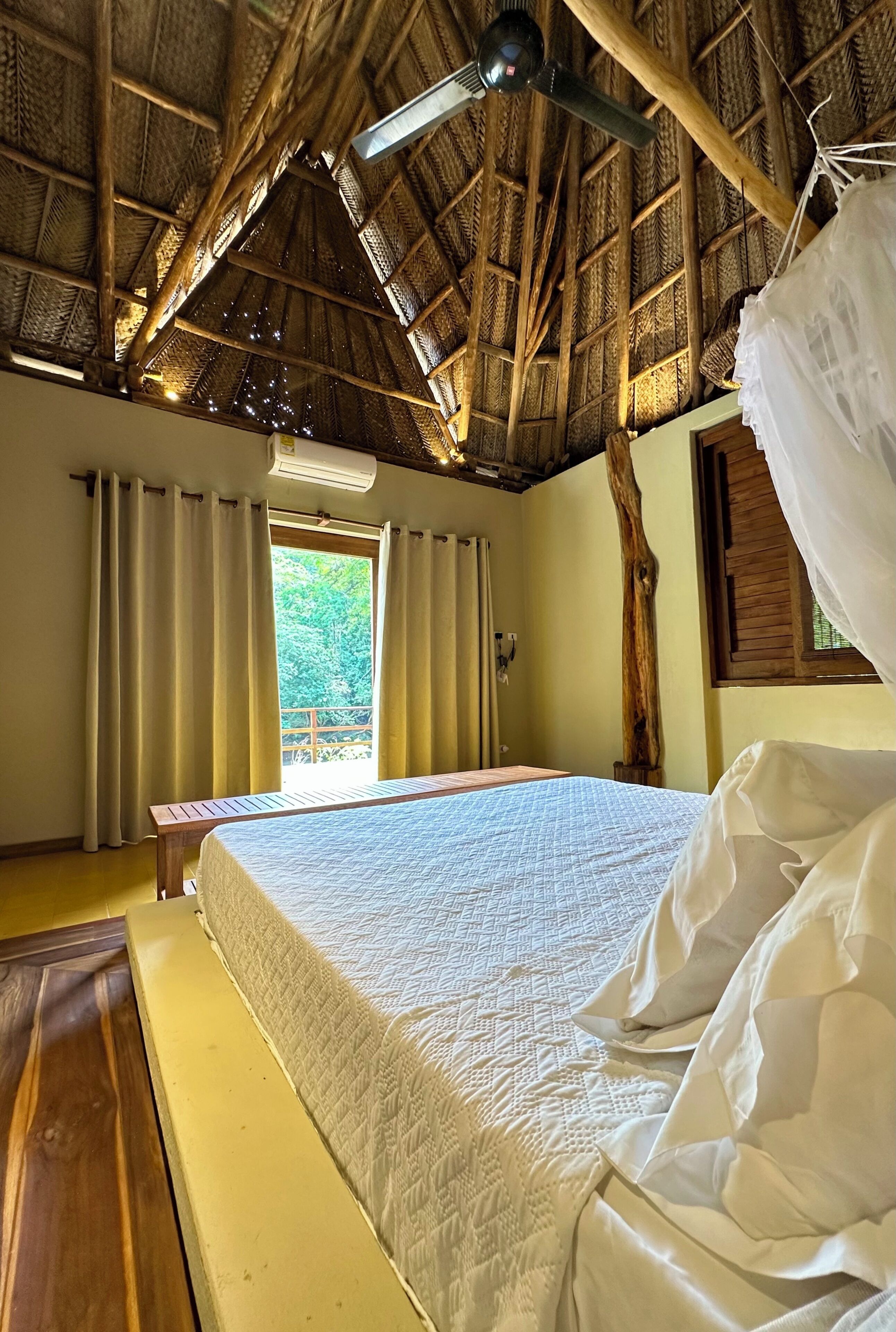 A room with a bed, a wooden bench, a window with curtains, a ceiling fan, and a view of greenery outside.