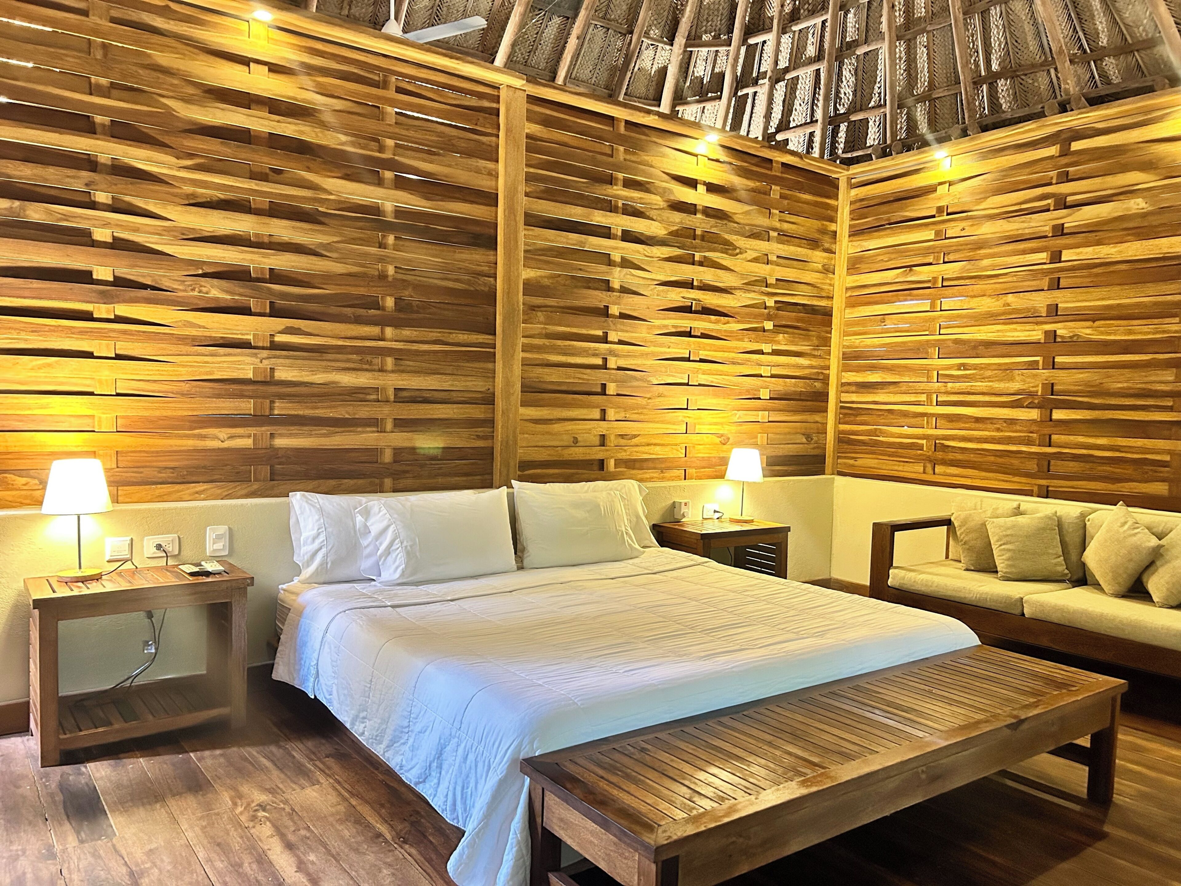 A room with a wooden slatted wall, a bed with white bedding, a wooden bench, and a sofa with cushions.