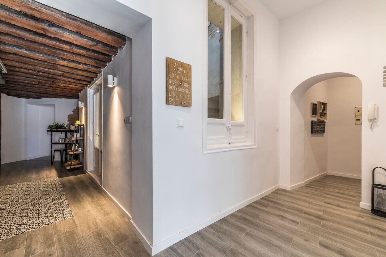 A hallway with a wooden ceiling, a window, and a door. There is a rug on the floor and a shelf with plants.