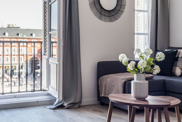 A modern living room with a large window, a grey sofa, a small round table, and a vase with white flowers.