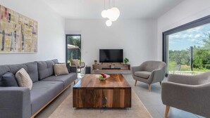 A modern living room with a grey sofa, a wooden coffee table, a TV on a wooden stand, and a large window offering a view of greenery. - Charming 4BR Villa with Private Pool and Nature Views (Vabriga)