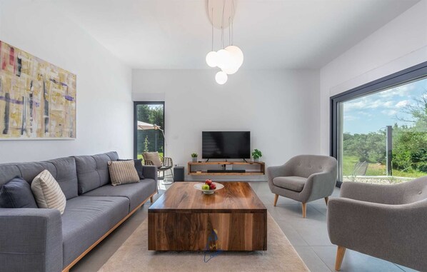 Un salon moderne avec un canapé gris, une table basse en bois, un téléviseur posé sur un meuble en bois et une grande fenêtre donnant sur un paysage verdoyant.