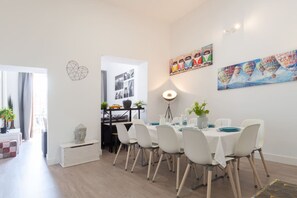 A modern dining area with white chairs and a round table, a wall-mounted light, and a painting of hot air balloons.