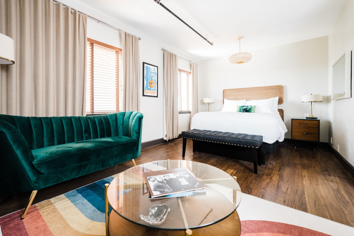 A hotel room with a bed, a green sofa, a glass coffee table, and a wooden floor.