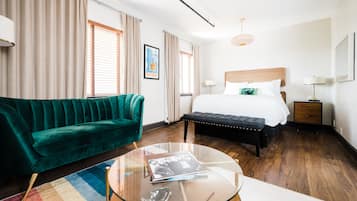 Quarto de hotel com uma cama, um sofá verde, uma mesa de centro de vidro e piso de madeira.