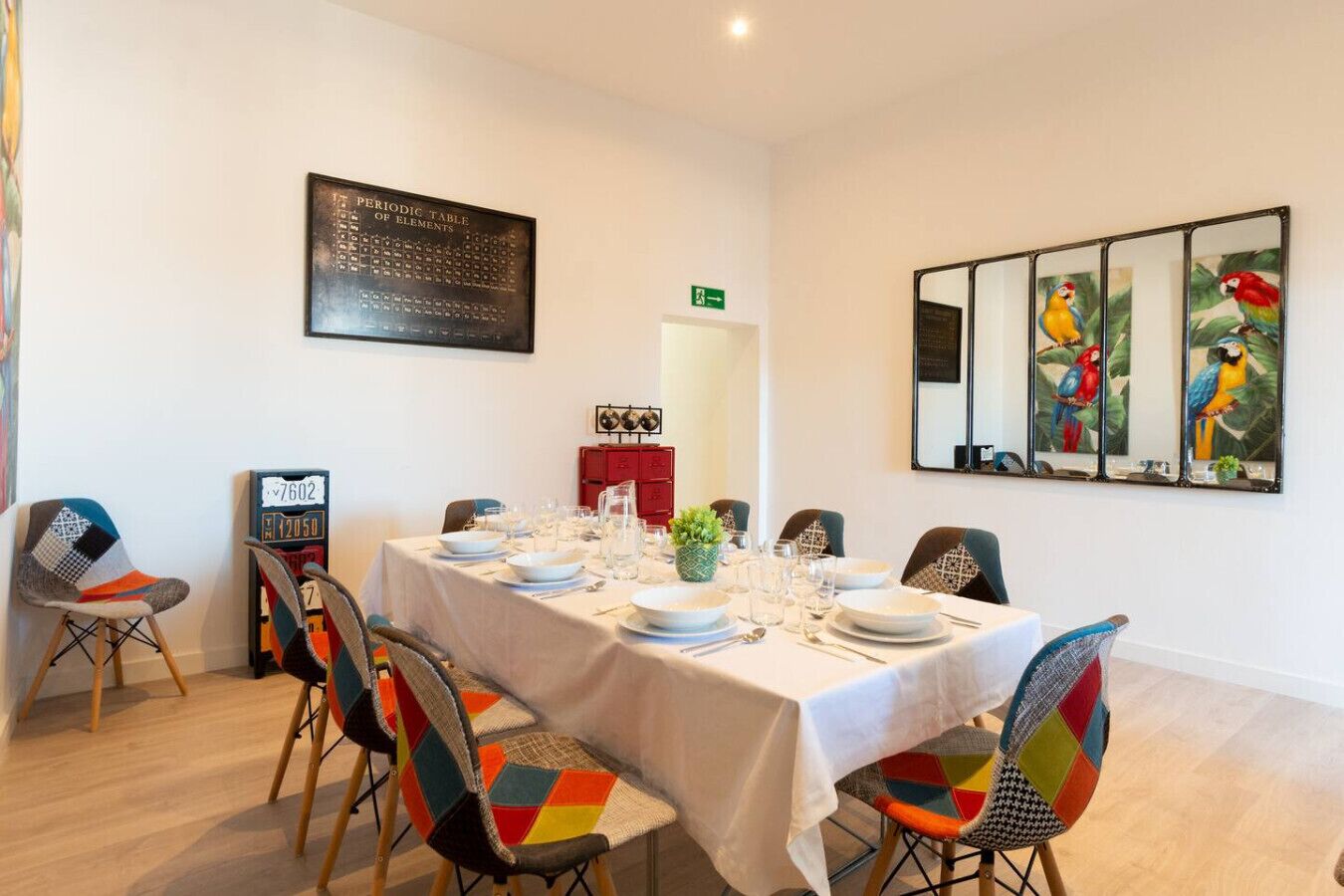 Comedor con una mesa larga dispuesta para seis personas, rodeada de sillas coloridas. Se exhibe una tabla periódica de elementos en la pared, y también se ve un cuadro de un loro colorido.