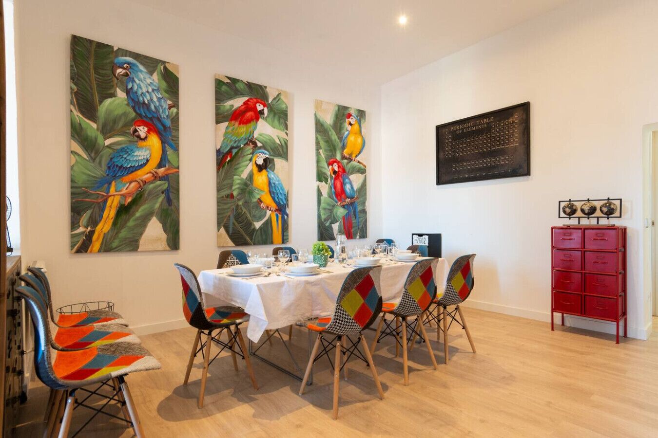 Un coin repas avec une table dressée pour quatre personnes, entourée de chaises aux couleurs vives, et trois grandes peintures représentant des perroquets accrochées au mur.