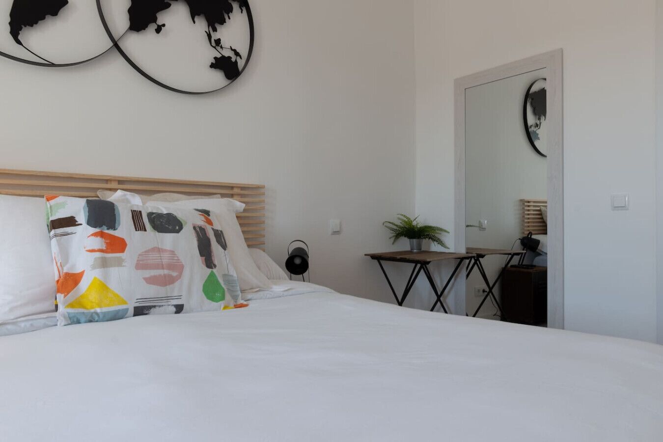 Ein ordentlich bezogenes Bett mit weißer Bettwäsche und bunten Kissen, ein kleiner Tisch mit einer Pflanze und ein an der Wand befestigter Spiegel.