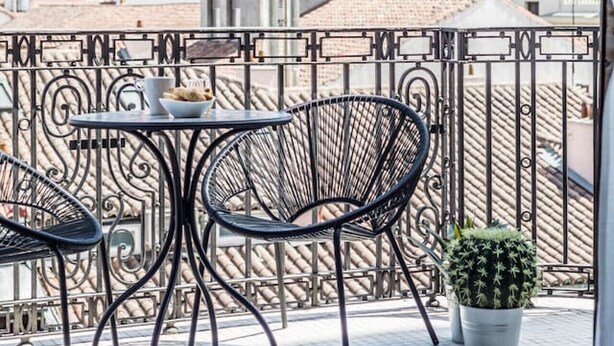 Een balkon met metalen stoelen en een tafel, een cactus in een pot, en een uitzicht op gebouwen op de achtergrond.