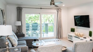 Una sala de estar moderna con un sofá gris, sillones estampados, un televisor y un balcón con mobiliario exterior.