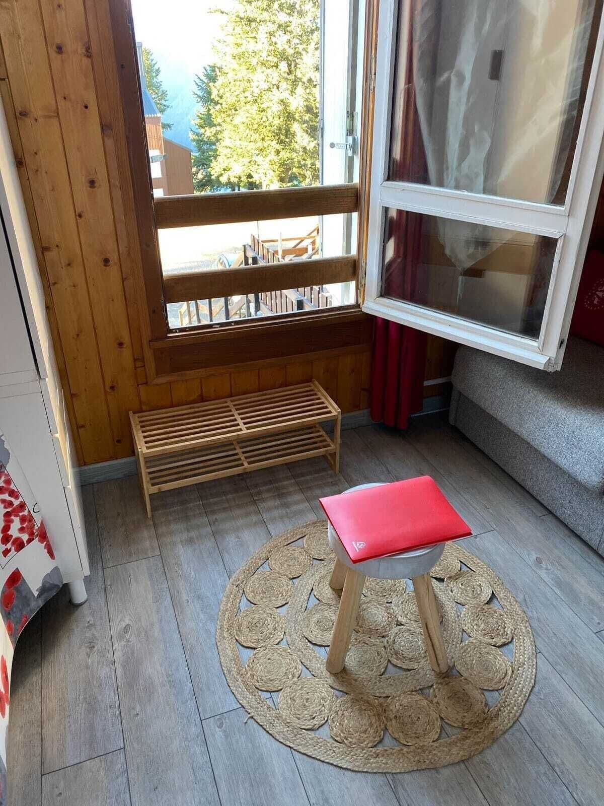 Une pièce avec un banc en bois, un petit tabouret, un tapis tissé et une fenêtre donnant sur des arbres et un bâtiment à l’extérieur.