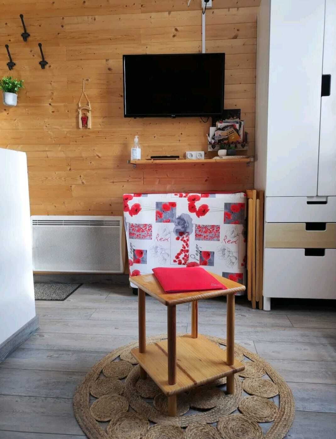 Una habitación moderna con una pared de madera, un televisor de pantalla plana montado sobre un estante, una pequeña mesa de madera con un cojín rojo y un mantel estampado.