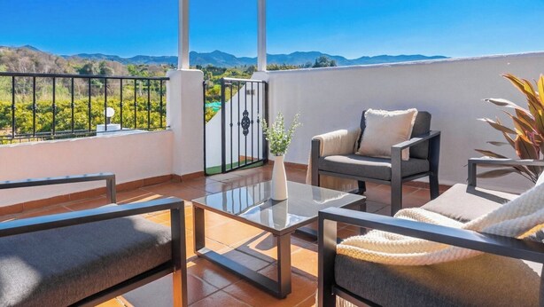 Un balcon avec une table en verre, deux fauteuils et une vue sur les montagnes et la verdure.