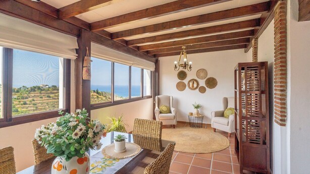 A room with a view of the sea, a dining area with wicker furniture, and a seating area with chairs and a round rug.