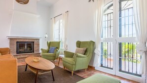 Una sala de estar con chimenea, sillones verdes, una mesa de centro de madera y ventanas grandes con cortinas.