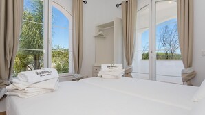 Habitación de hotel con una cama grande, dos ventanas con cortinas, vistas a espacios verdes y una pila de toallas blancas con un logotipo.