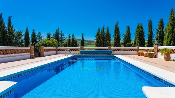 Piscina rectangular con superficie de agua azul cristalina, rodeada por un borde de baldosas blancas y un muro de ladrillo bajo.