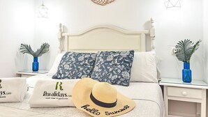 Ein ordentlich gemachtes Bett mit blumigen Kissen, einem Strohhut und einer blauen Vase mit Pflanze auf einem Nachttisch.