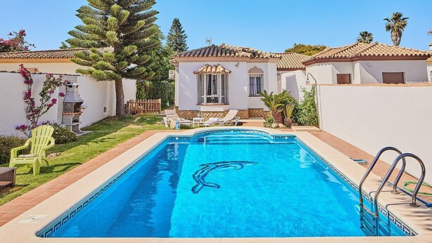 Ein rechteckiger Swimmingpool mit klarem, blauem Wasser, umgeben von einer gefliesten Liegefläche und einem kleinen Gartenbereich mit Stühlen und einem Tisch.