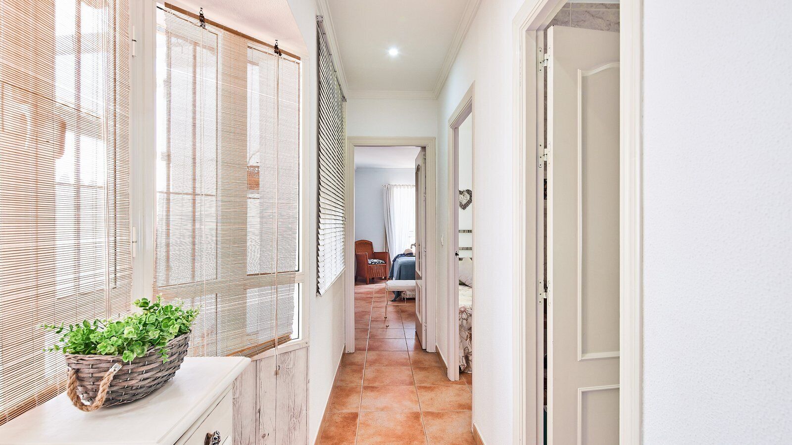A hallway with a wicker basket of green plants, wooden blinds, and a tiled floor leading to a bedroom with a bed and a chair.