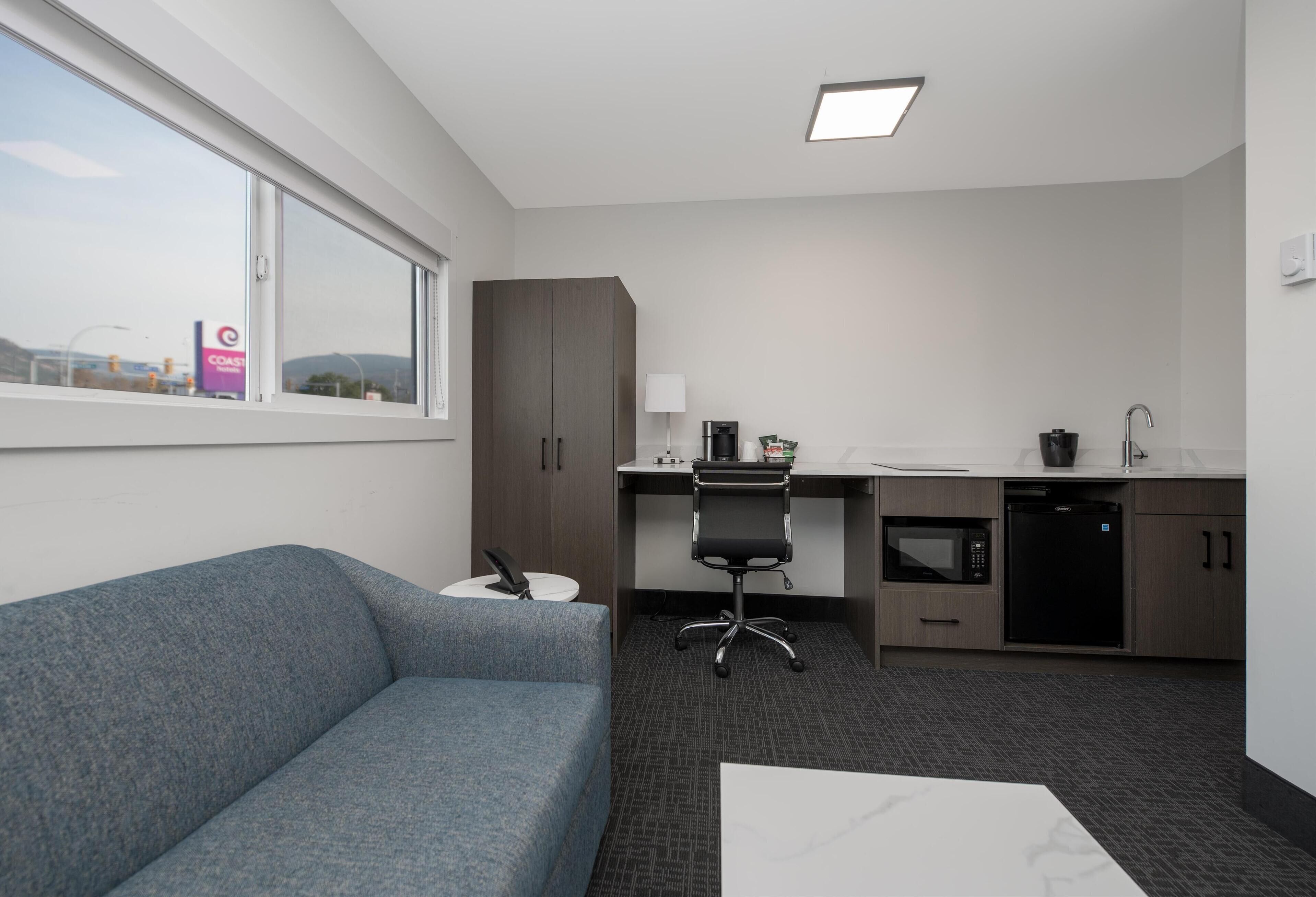 Una habitación de hotel moderna con un sofá gris, una mesa de centro blanca, un escritorio con una silla, un microondas, un lavavajillas y una ventana con vistas a un edificio lejano.
