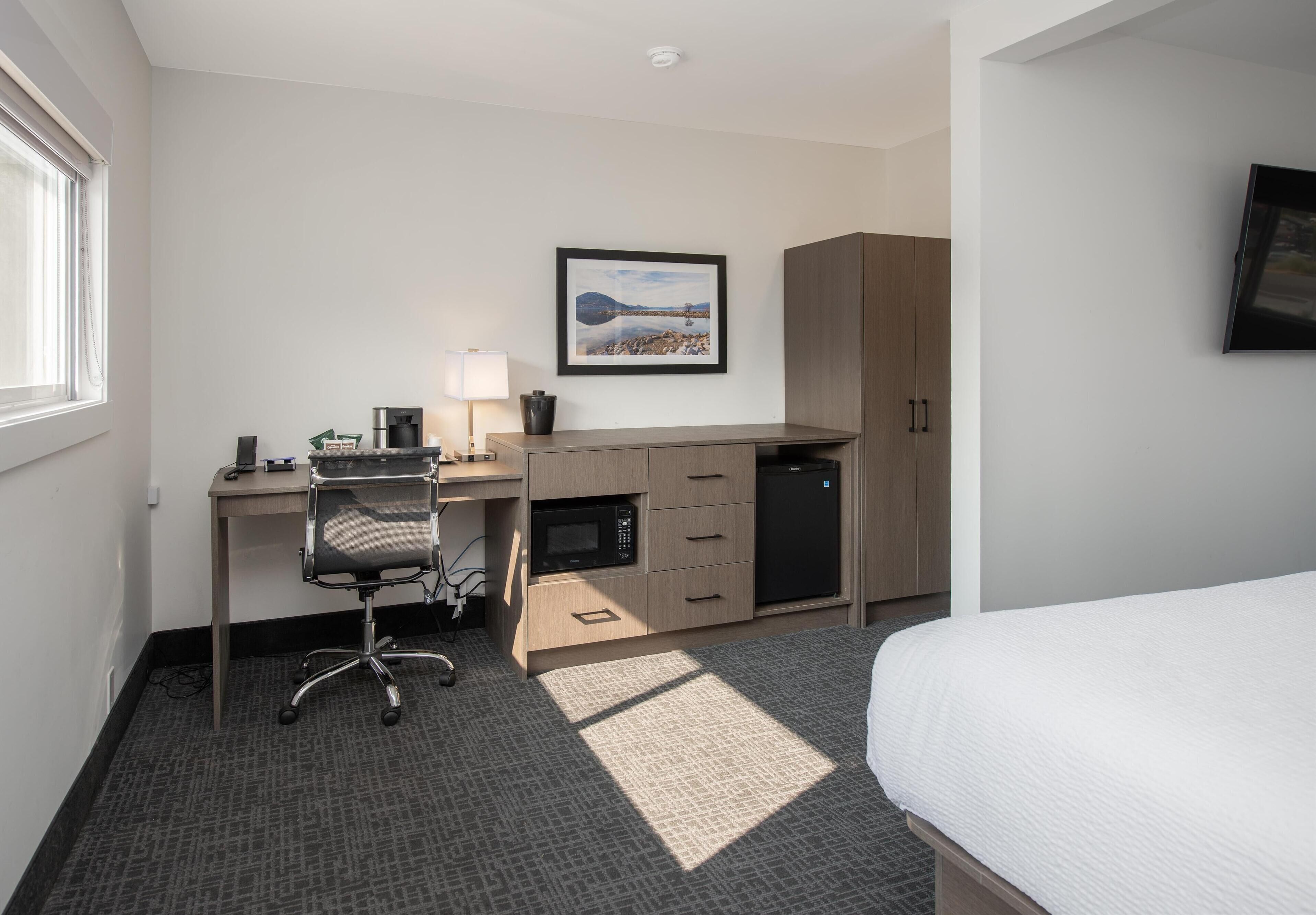 Habitación de hotel con una cama, un escritorio con computadora, un microondas, cafetera, televisor y un cuadro en la pared.