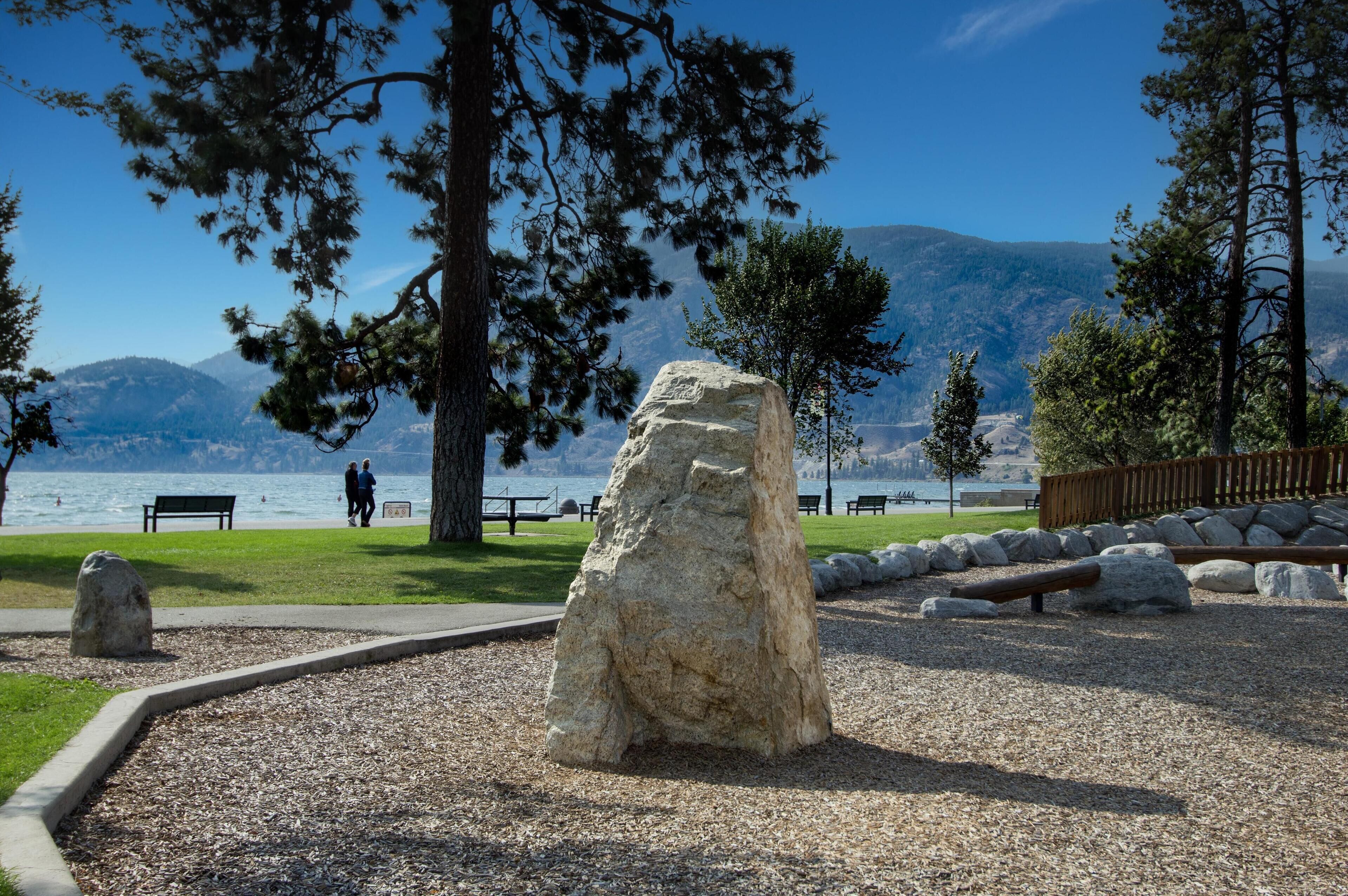 Un parque con una roca grande, bancos y vista a un lago.