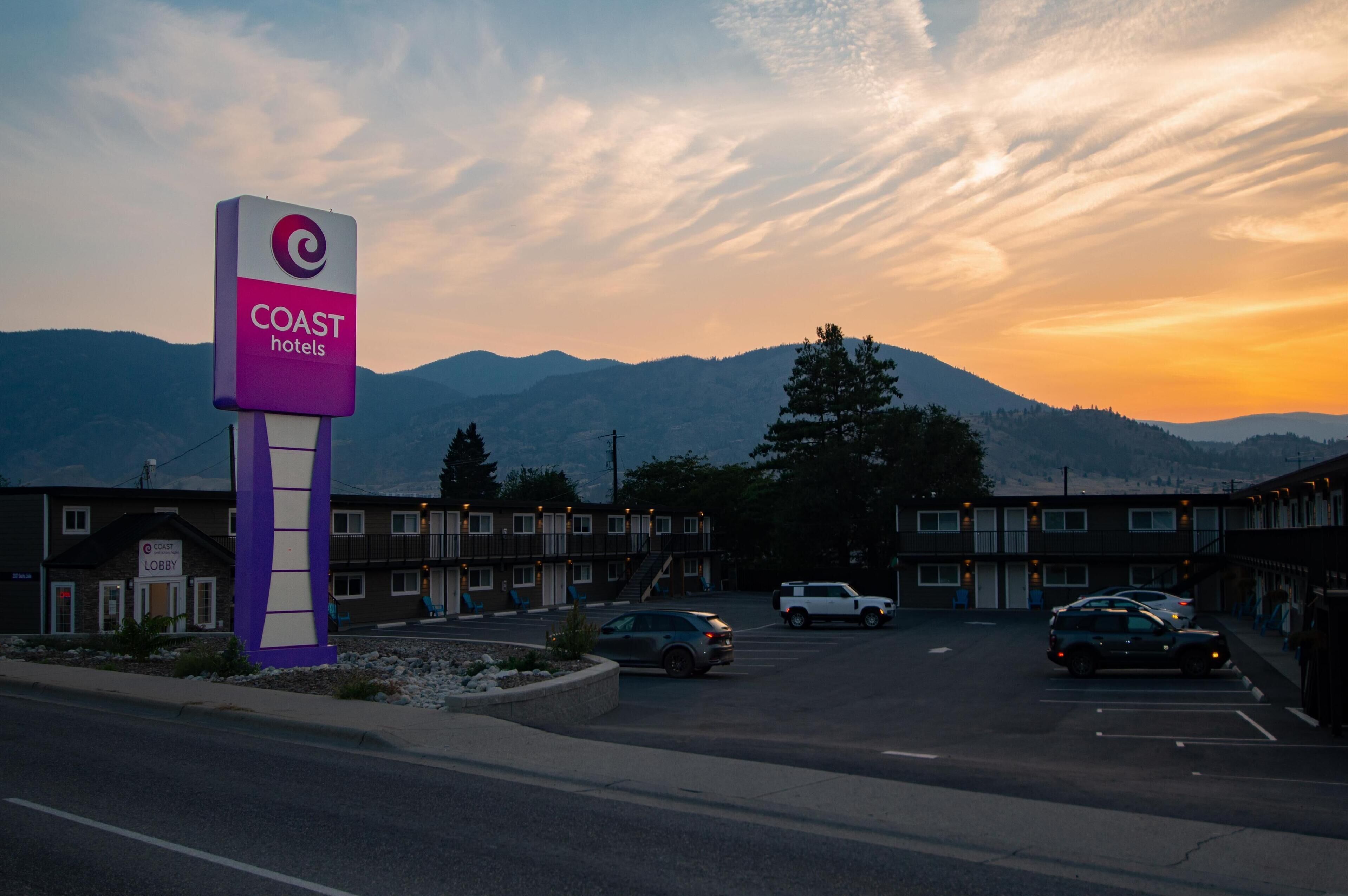 Un motel con un letrero llamativo, varios espacios de estacionamiento y montañas al fondo.
