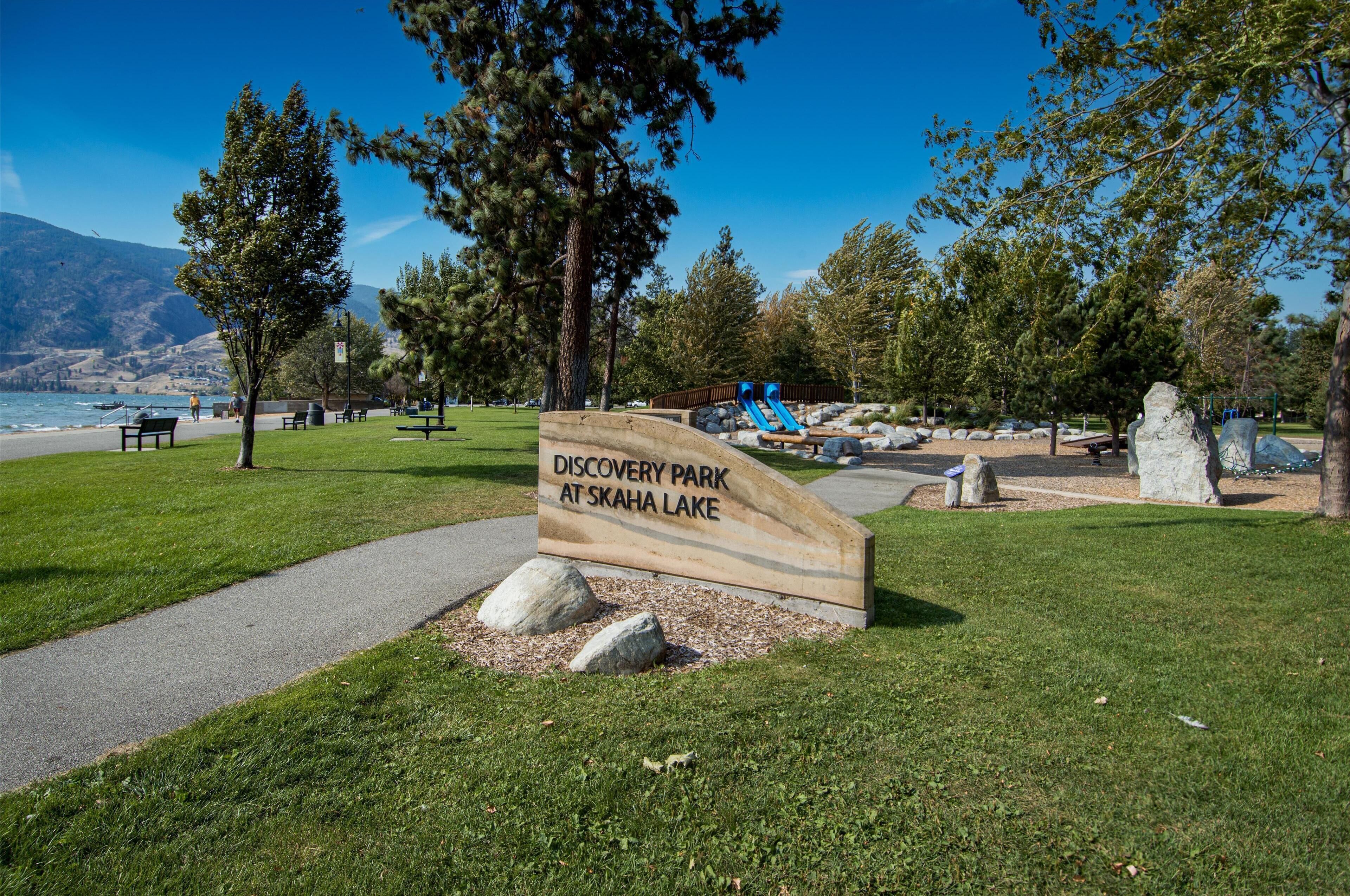 A park sign for Discovery Park at Skaha Lake with a path, benches, and a playground.