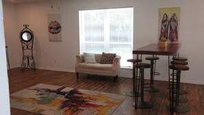 A living room with a sofa, a coffee table, a bar stool set, a colorful rug, a wall clock, and a window with blinds.