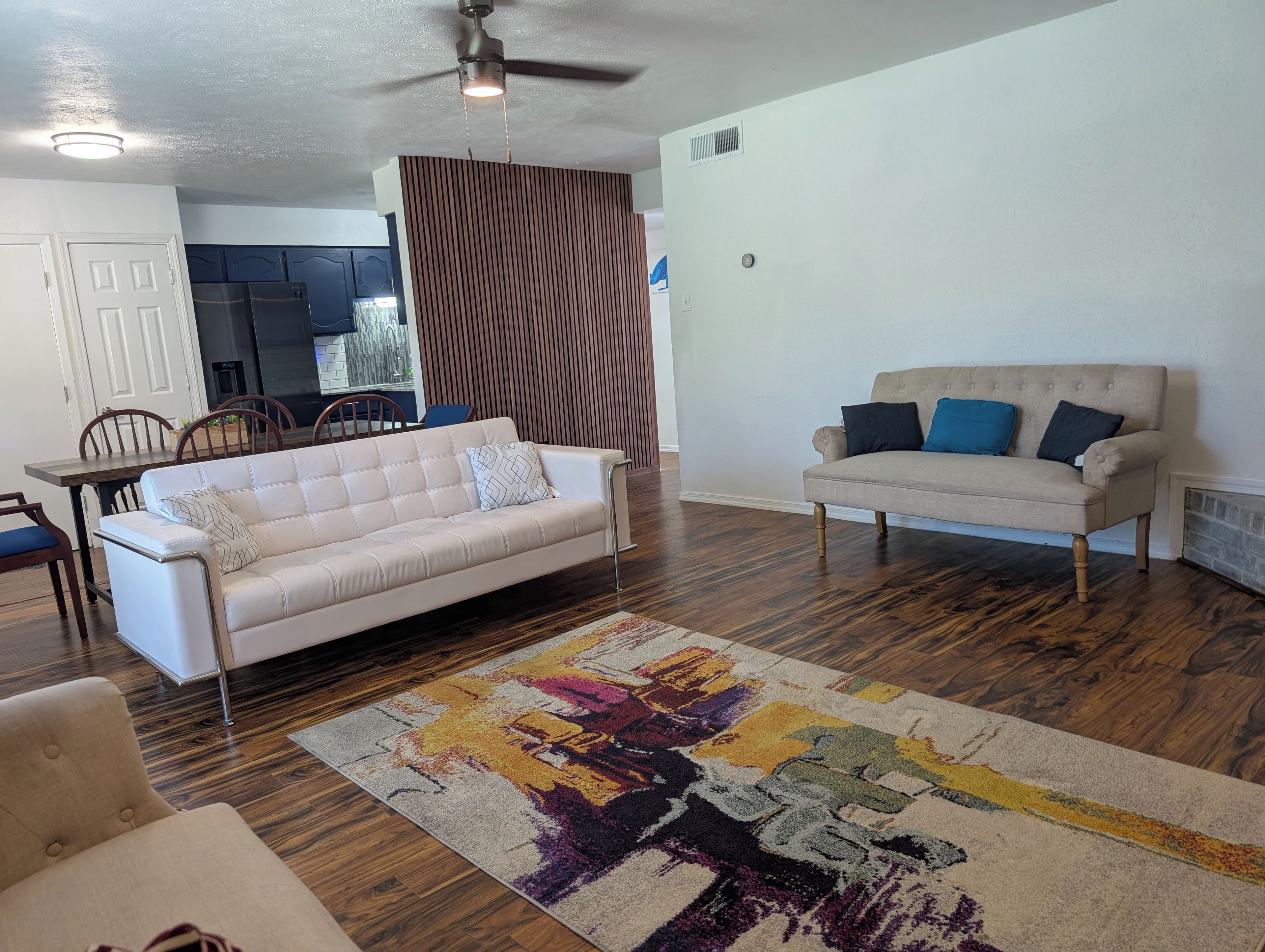 A living room with a white sofa, a beige sofa, a patterned rug, wooden flooring, a ceiling fan, and a kitchen in the background.